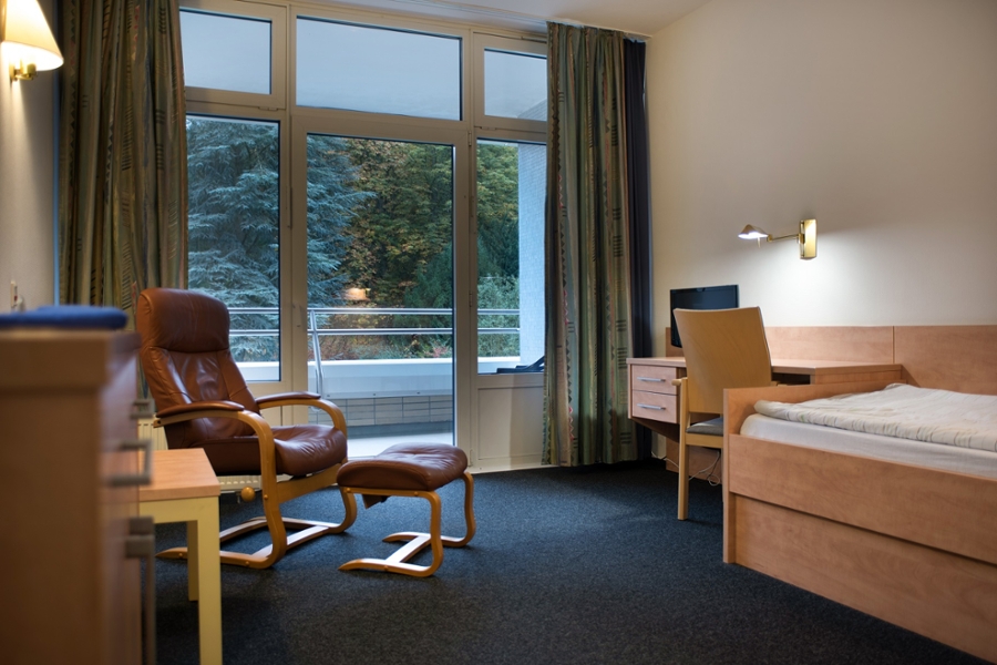 Patientenzimmer mit einem Liegesessel, einem Fernseher auf einem Schreibtisch nebst dazugehörigem Stuhl sowie einem Bett mit Blick auf die parkähnlichen Grünanlagen.