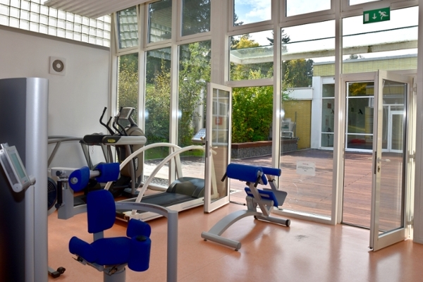 Ein Wintergarten, in dem verschiedene sporttherapeutische Geräte stehen. An die Fensterscheiben grenzen eine holzgetäfelte Terrasse und Grünpflanzen.